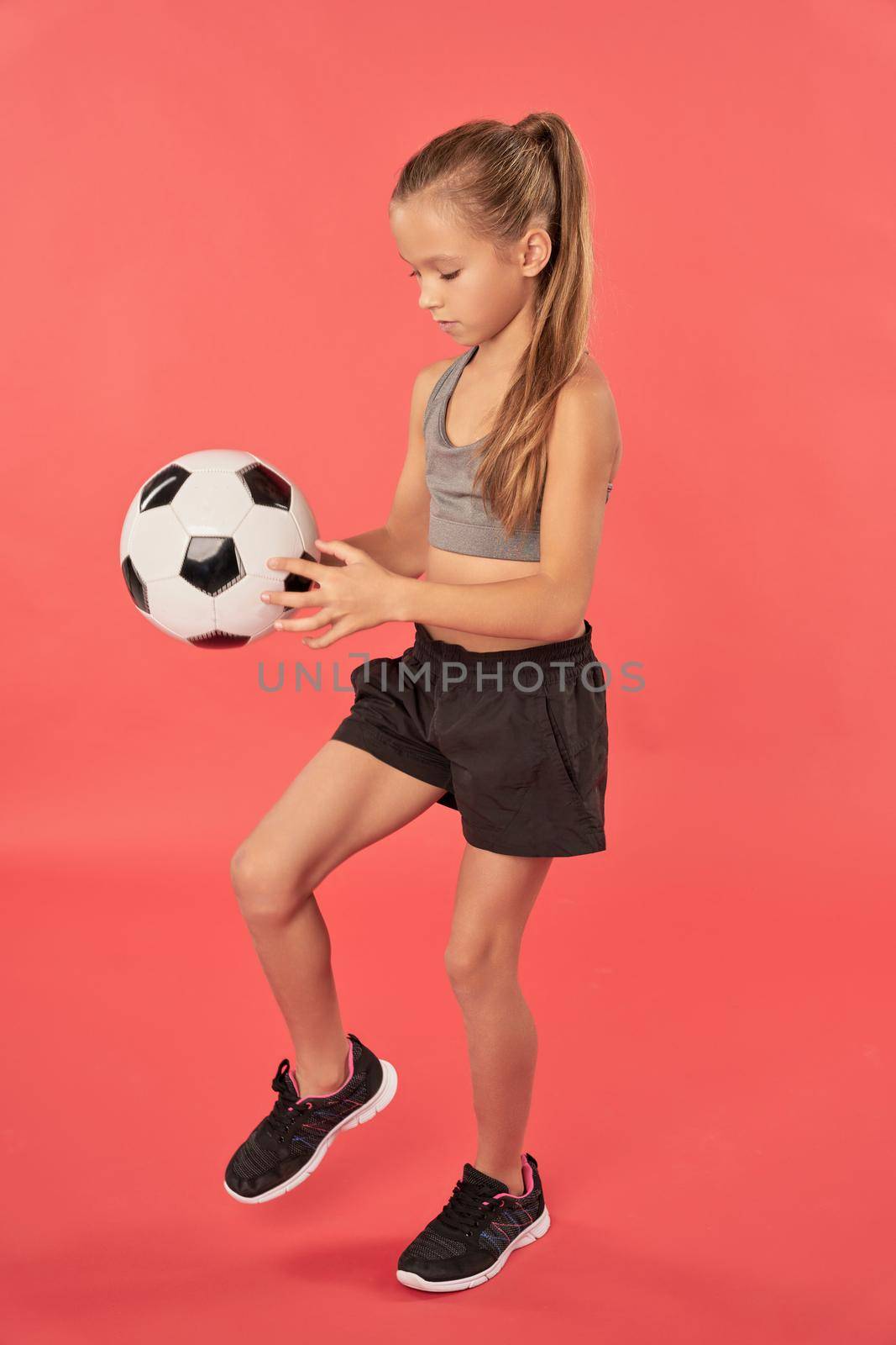 Cute female child in sportswear kicking football ball with knee while standing against red background