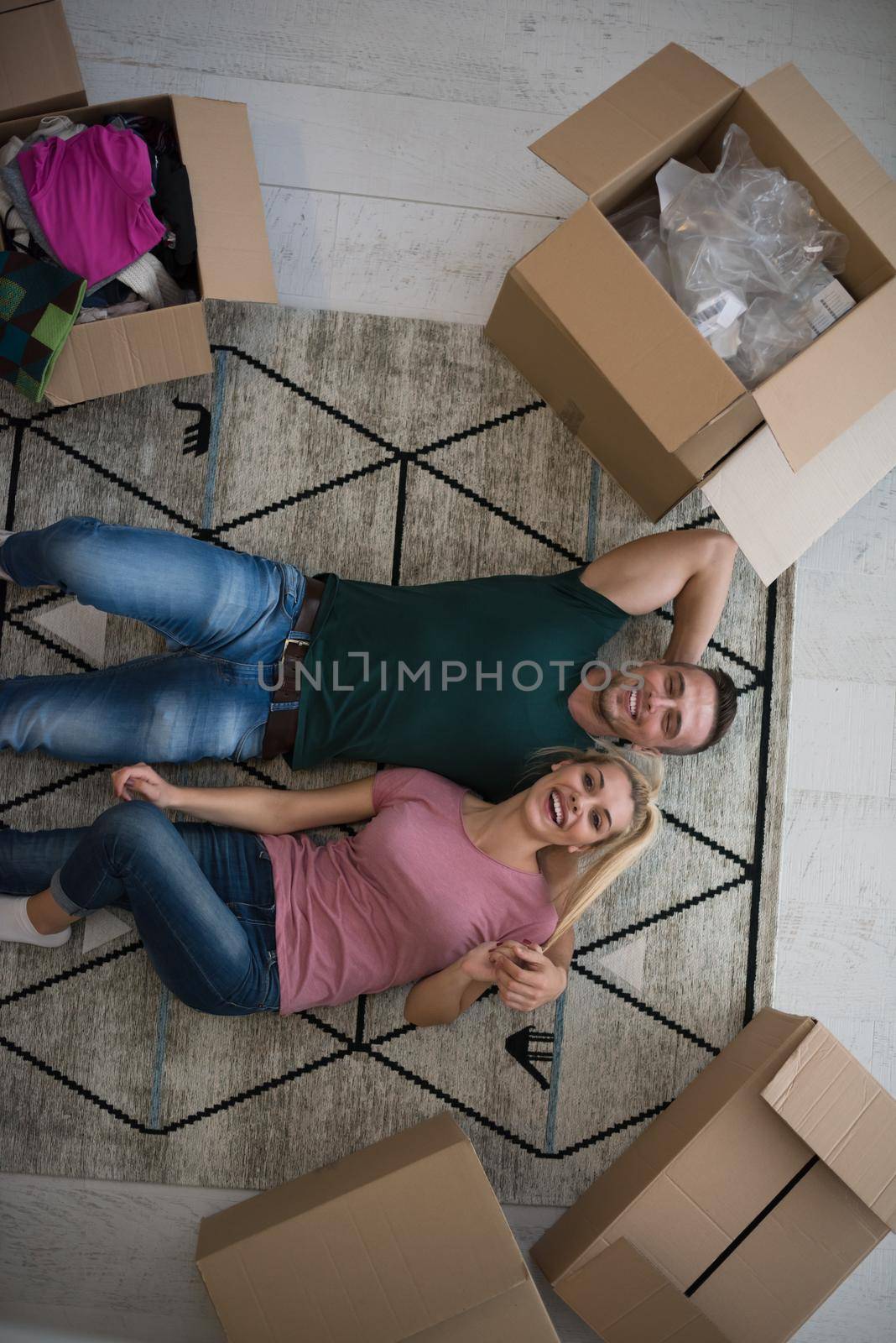 Top view of attractive young couple moving, holding hands, looking at camera and smiling while lying among cardboard boxes