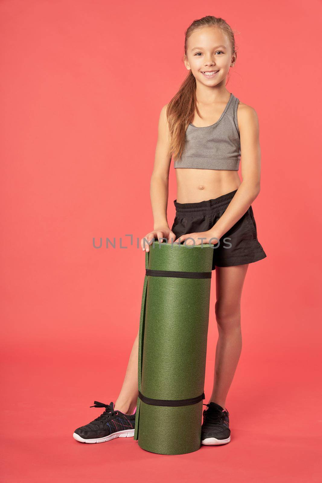 Adorable female child in sportswear looking at camera and smiling while holding rolled exercise mat