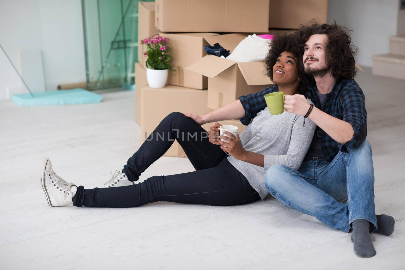 Relaxing in new house. Cheerful young multiethnic couple sitting on the floor and drinking coffee while cardboard boxes laying all around them