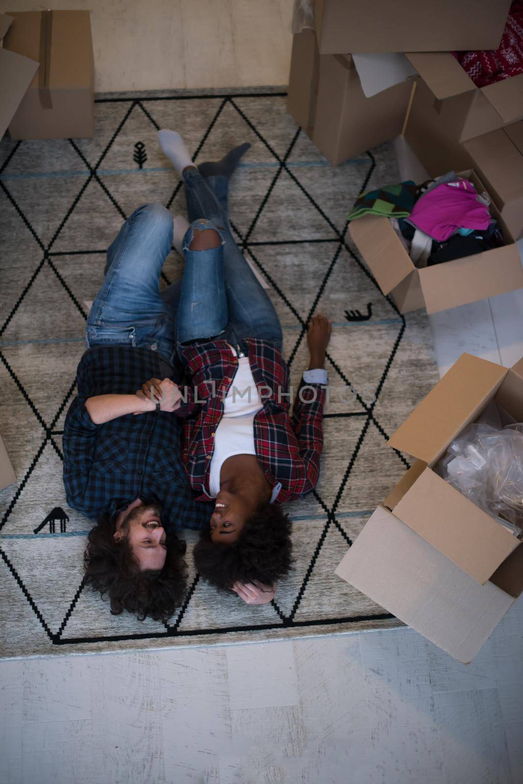 Top view of attractive young multiethnic couple moving, holding hands, looking at camera and smiling while lying among cardboard boxes