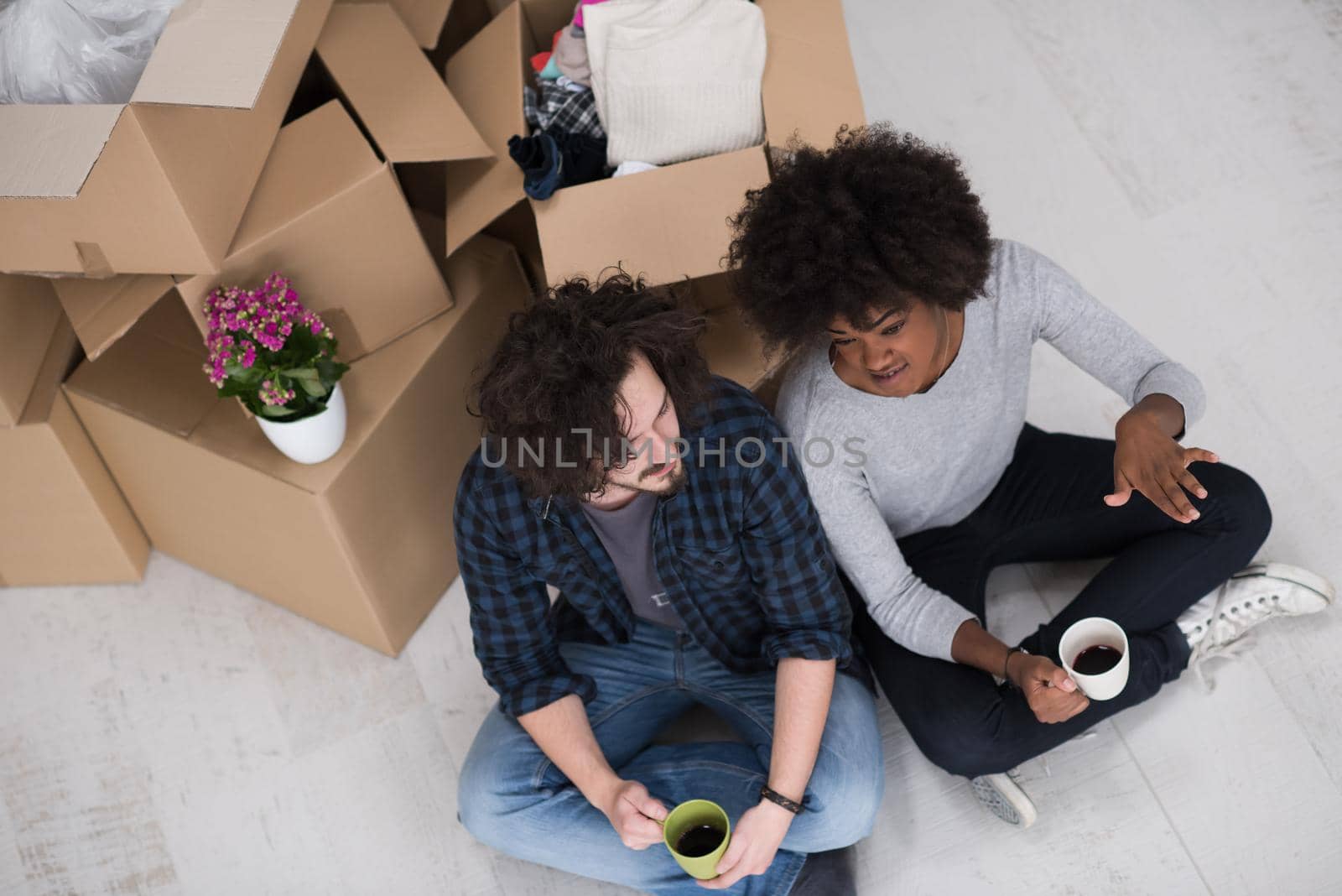 Relaxing in new house. Cheerful young multiethnic couple sitting on the floor and drinking coffee while cardboard boxes laying all around them