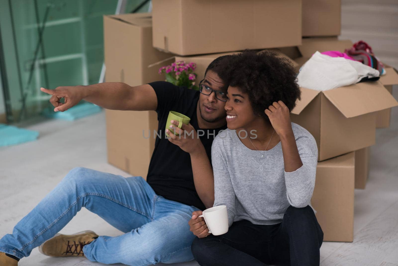 Relaxing in new house. Cheerful young African American couple sitting on the floor and drinking coffee while cardboard boxes laying all around them