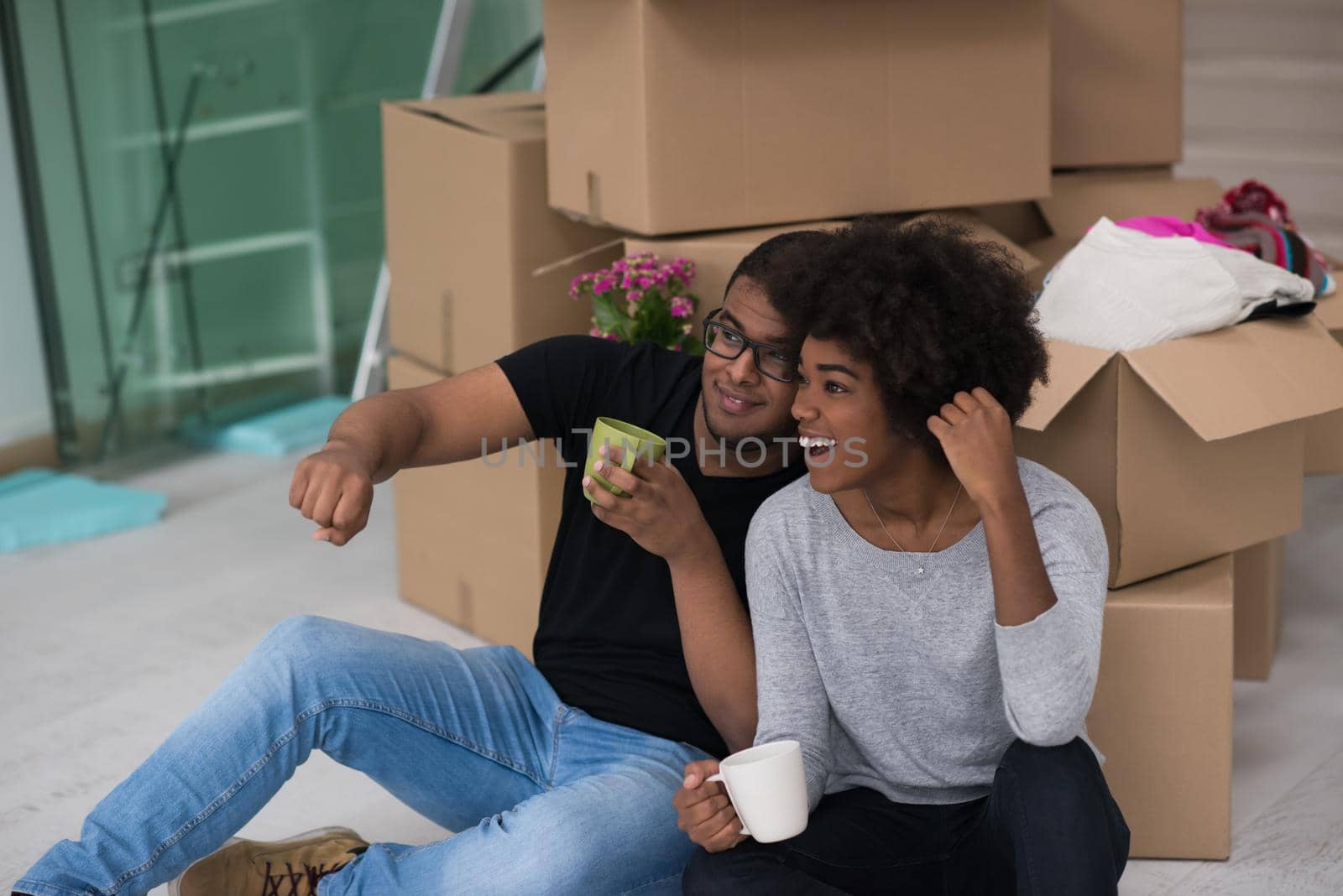 Relaxing in new house. Cheerful young African American couple sitting on the floor and drinking coffee while cardboard boxes laying all around them