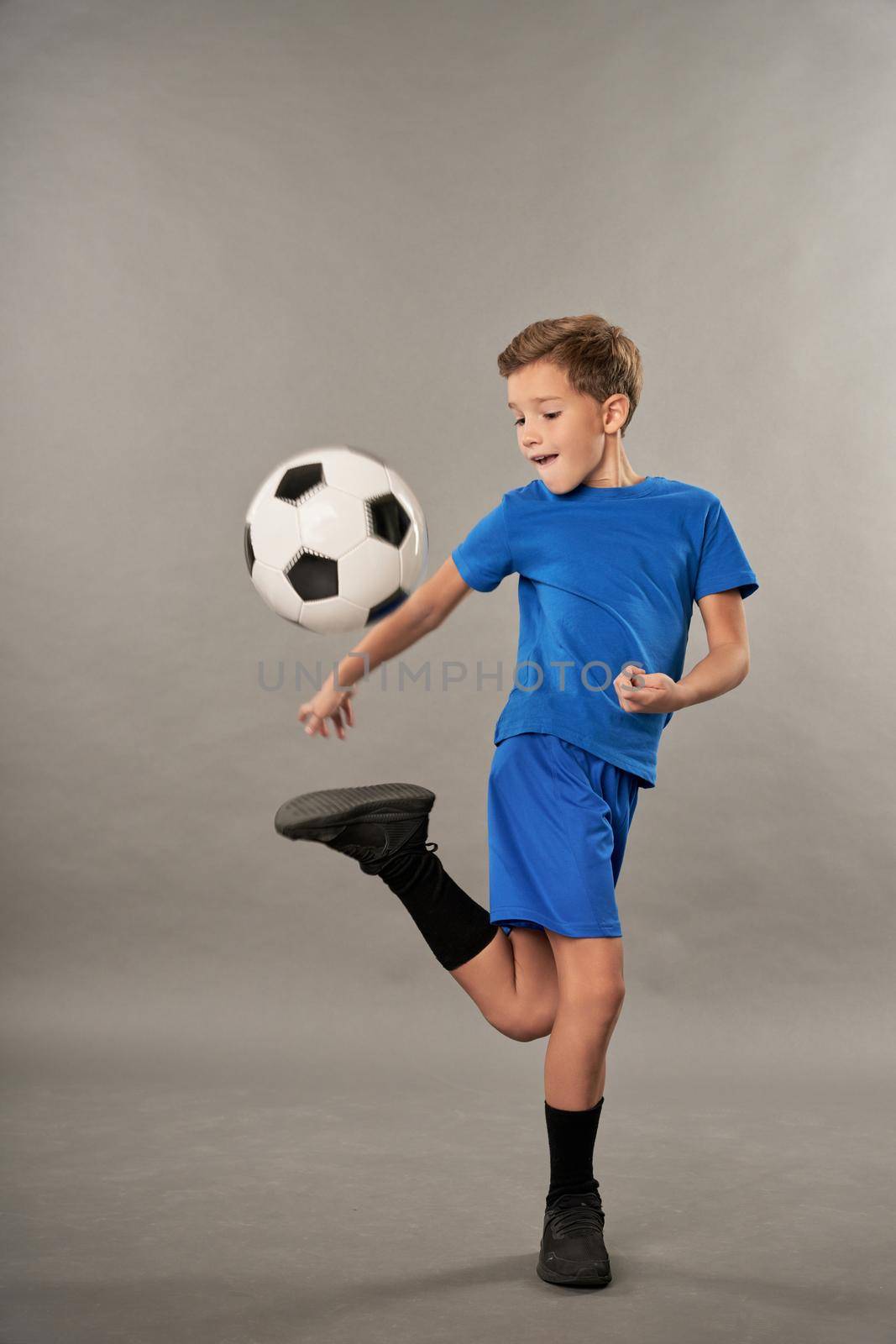 Adorable male child in shorts doing football tricks against gray background