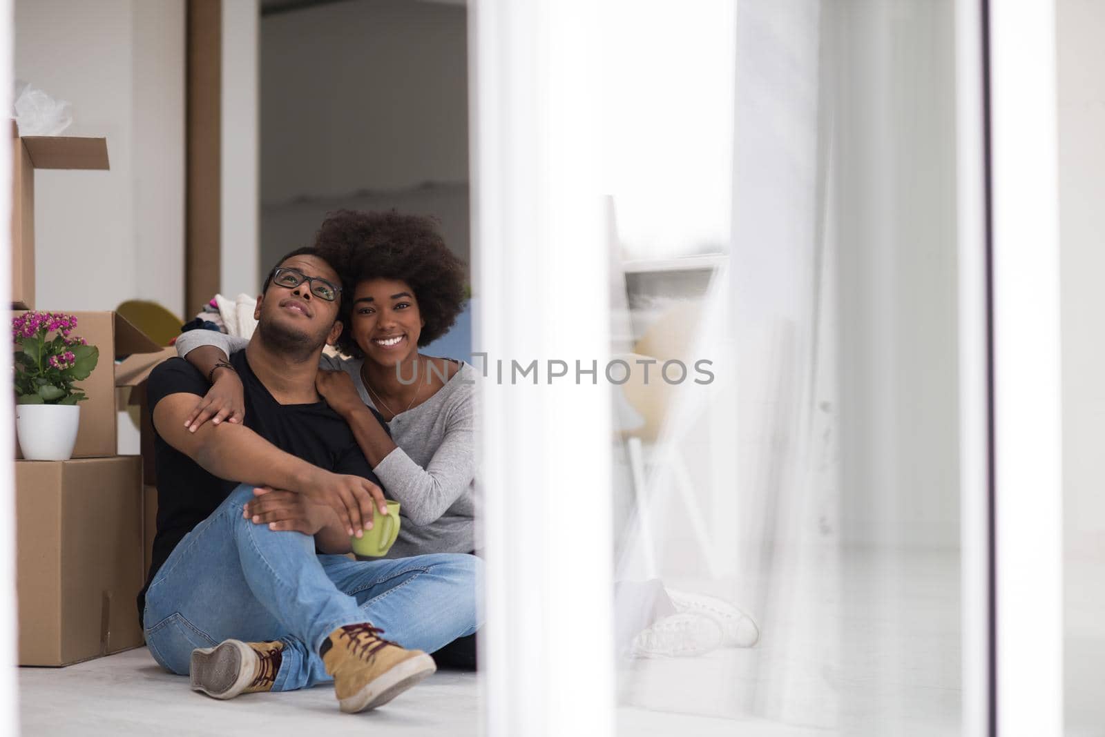 Relaxing in new house. Cheerful young African American couple sitting on the floor and drinking coffee while cardboard boxes laying all around them