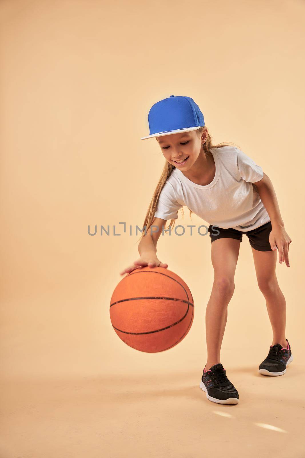 Adorable female child basketball player bouncing the ball and smiling while standing against light orange background