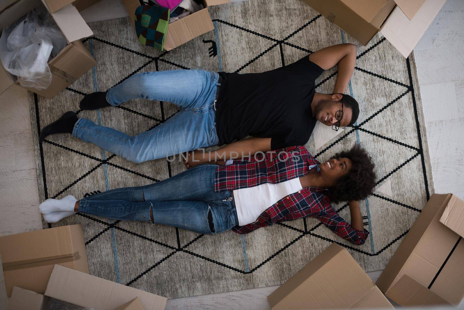 Top view of attractive young African American couple moving, holding hands, looking at camera and smiling while lying among cardboard boxes