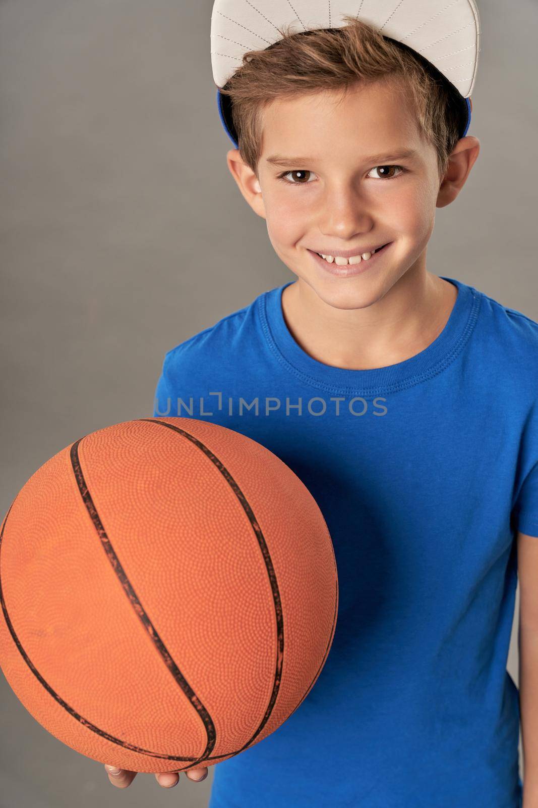 Adorable boy basketball player holding game ball and smiling while standing against gray background