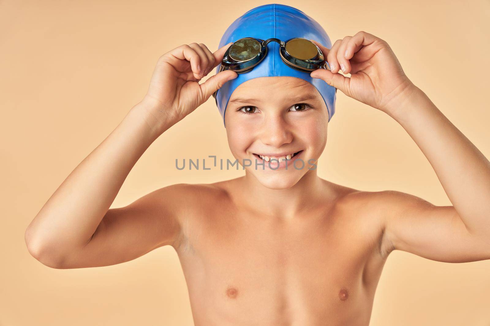 Close up of cheerful male child in swim cap looking at camera and smiling while adjusting swim goggles