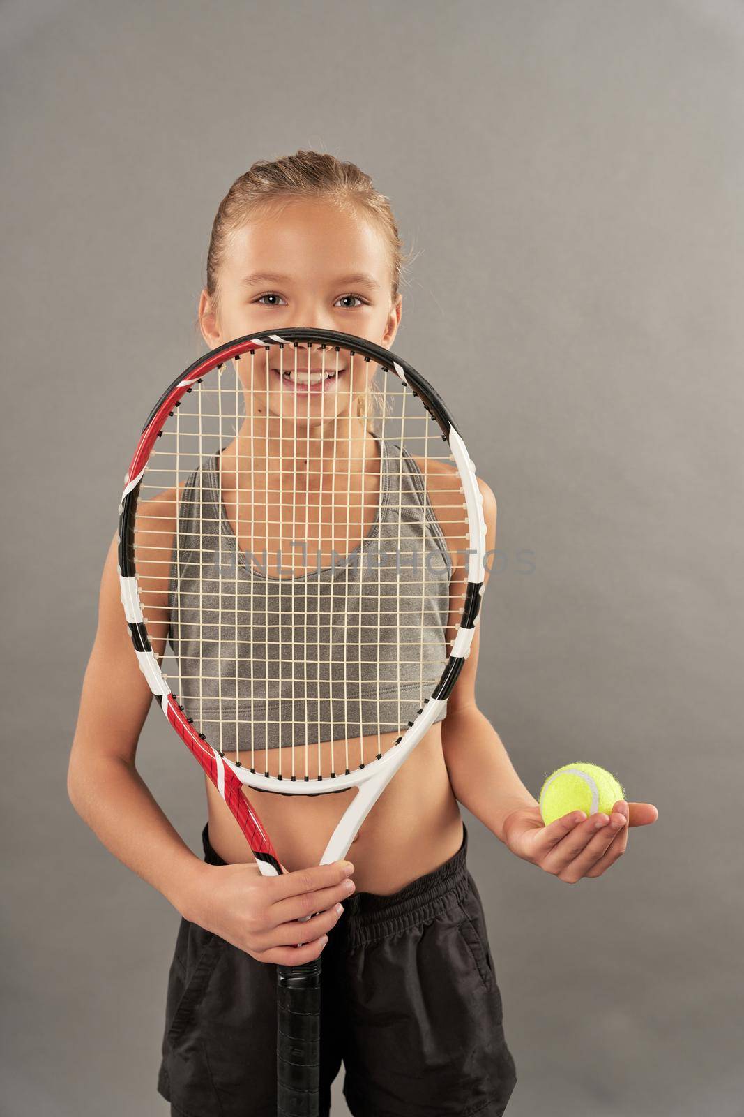 Cute girl tennis player covering face with racket and smiling while standing against gray background