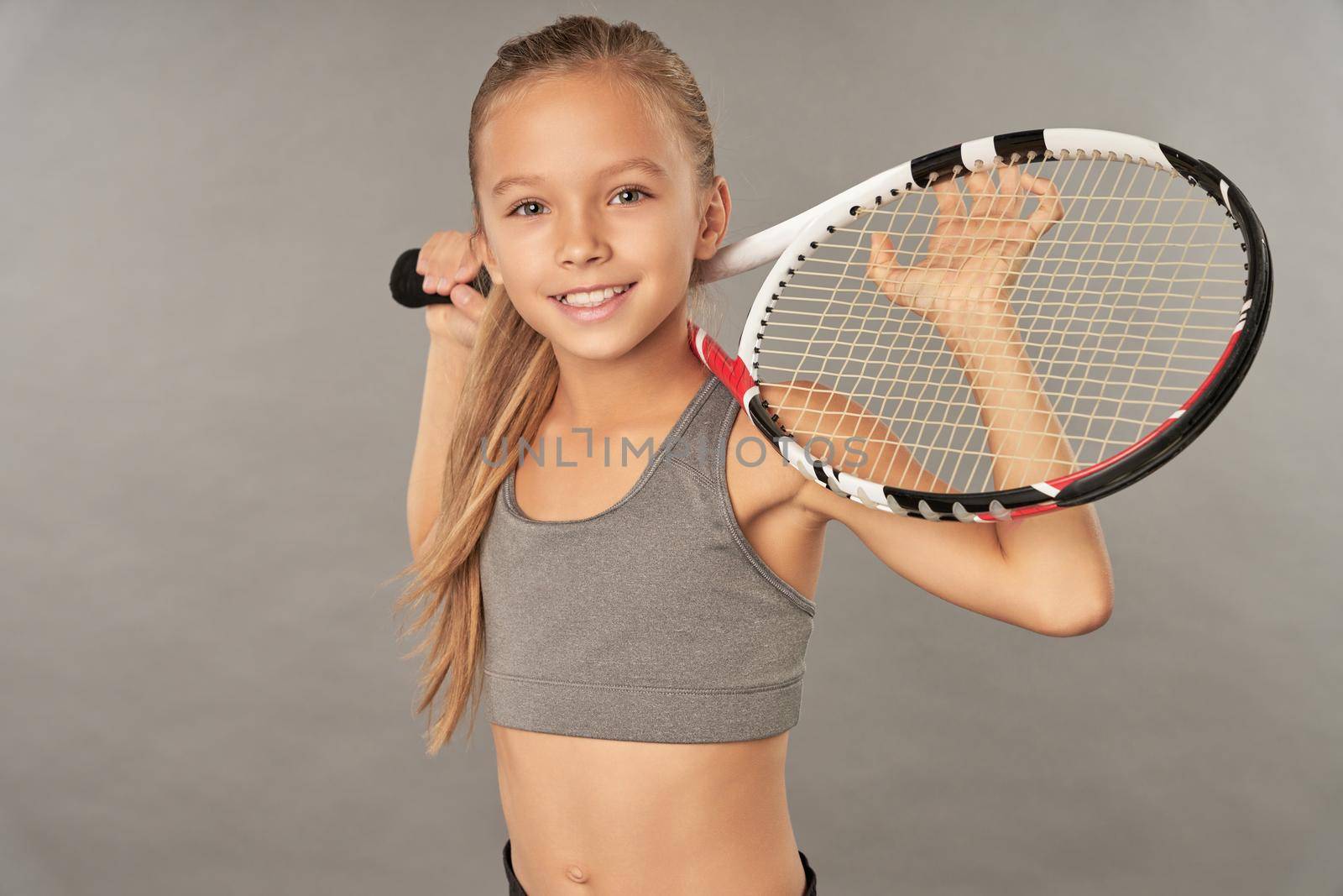 Cheerful female child tennis player looking at camera and smiling while holding racket behind her back