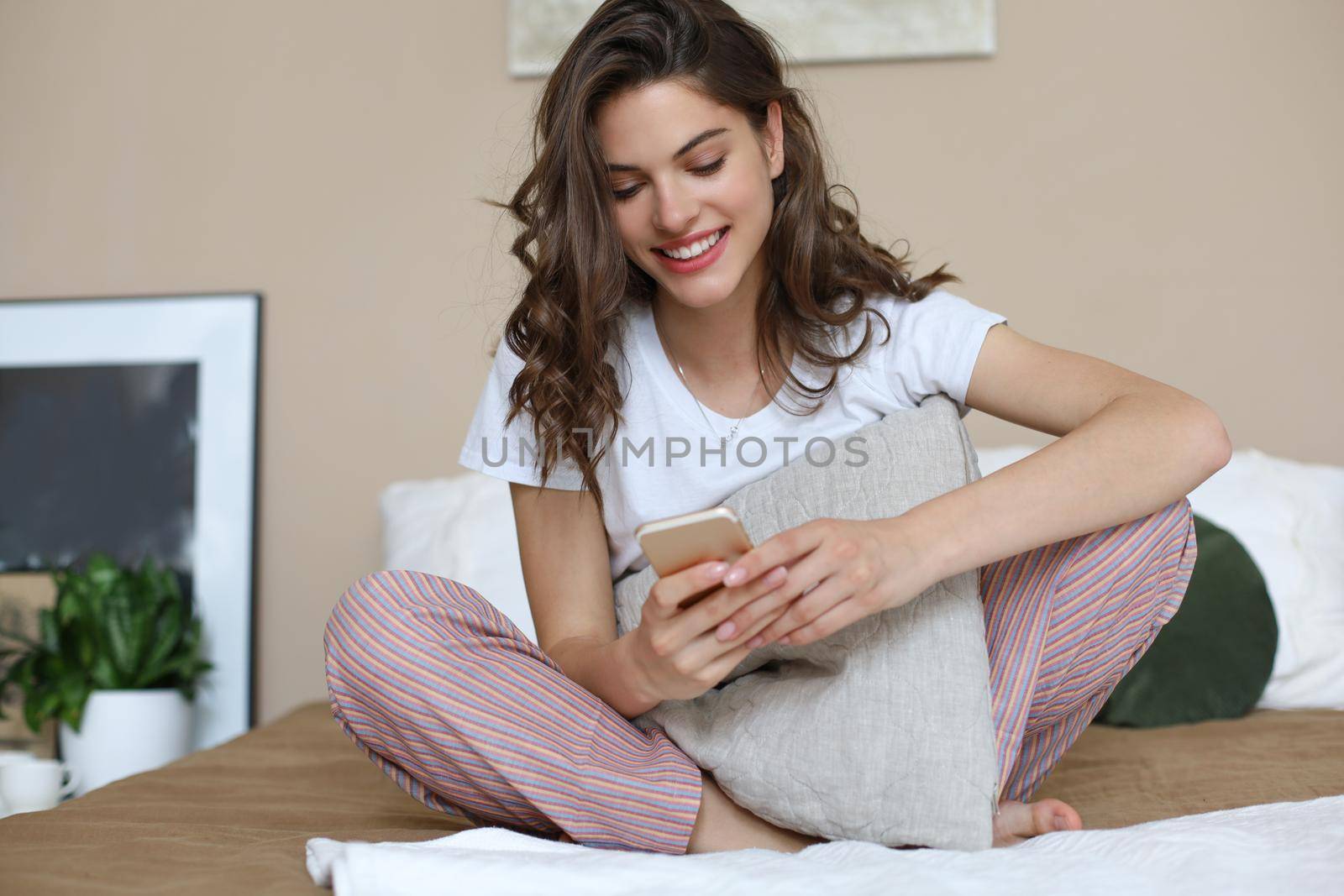 Beautiful young smiling brunette woman sitting in bed and using a phone in her bedroom