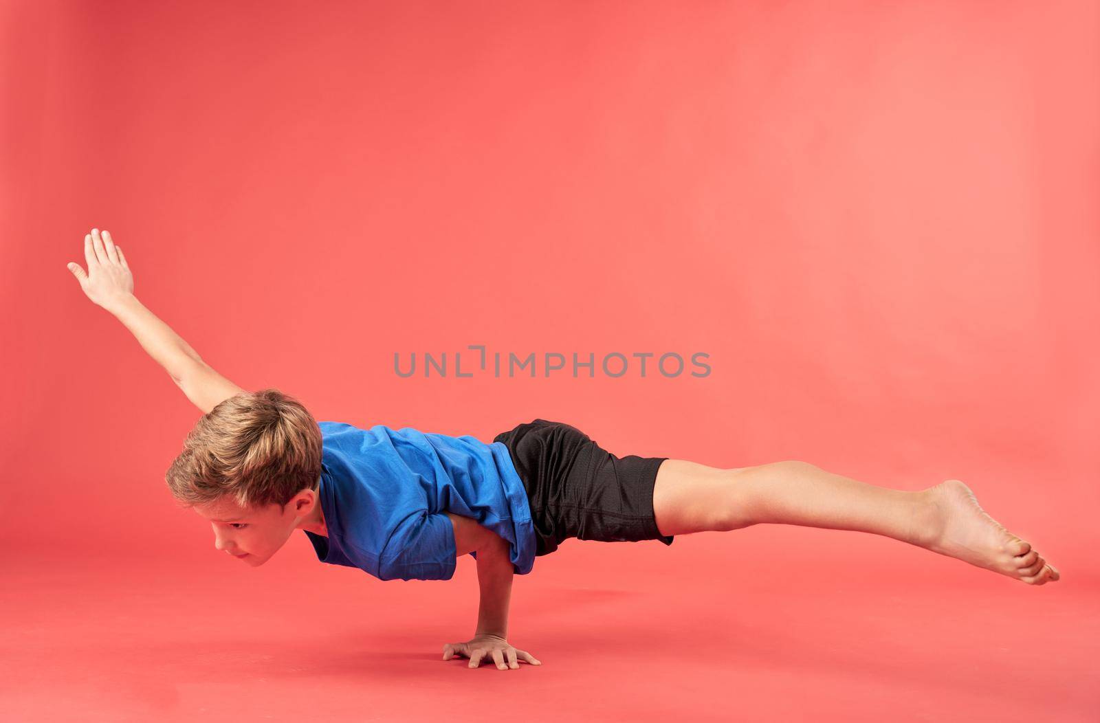 Cute male child in shorts balancing on one hand and looking away. Isolated on red background