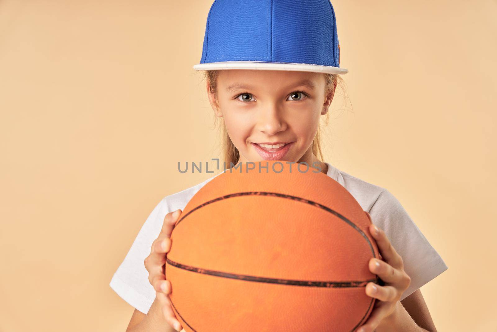 Cheerful female child basketball player holding game ball and smiling while standing against light orange background