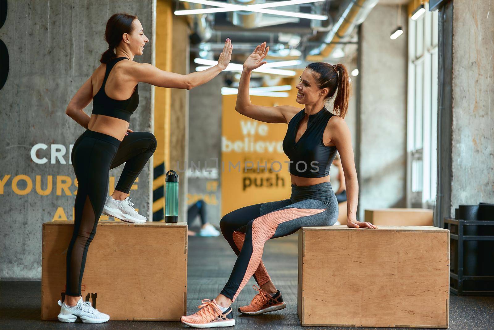 Great workout. Two young athletic fitness girls in sportswear giving high five each other while sitting on crossfit jump boxes at gym, exercising together. Sport, training and healthy lifestyle