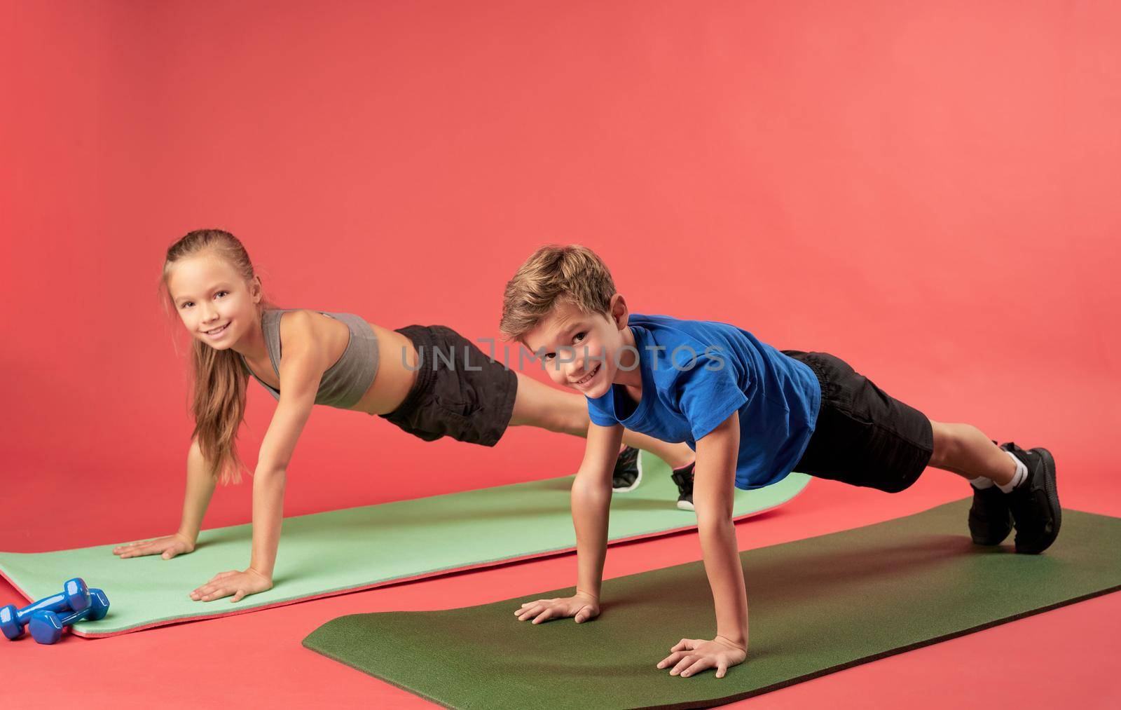 Adorable girl and boy holding push-up position and smiling while doing strength exercise on yoga mats