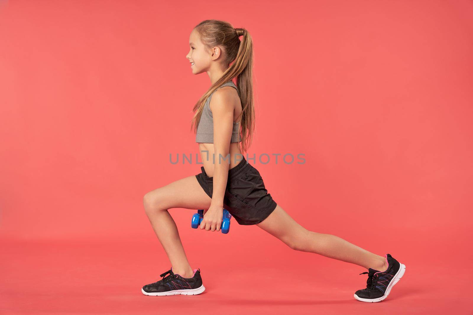 Cheerful female child in sportswear holding dumbbells and smiling while having morning workout. Isolated on red background