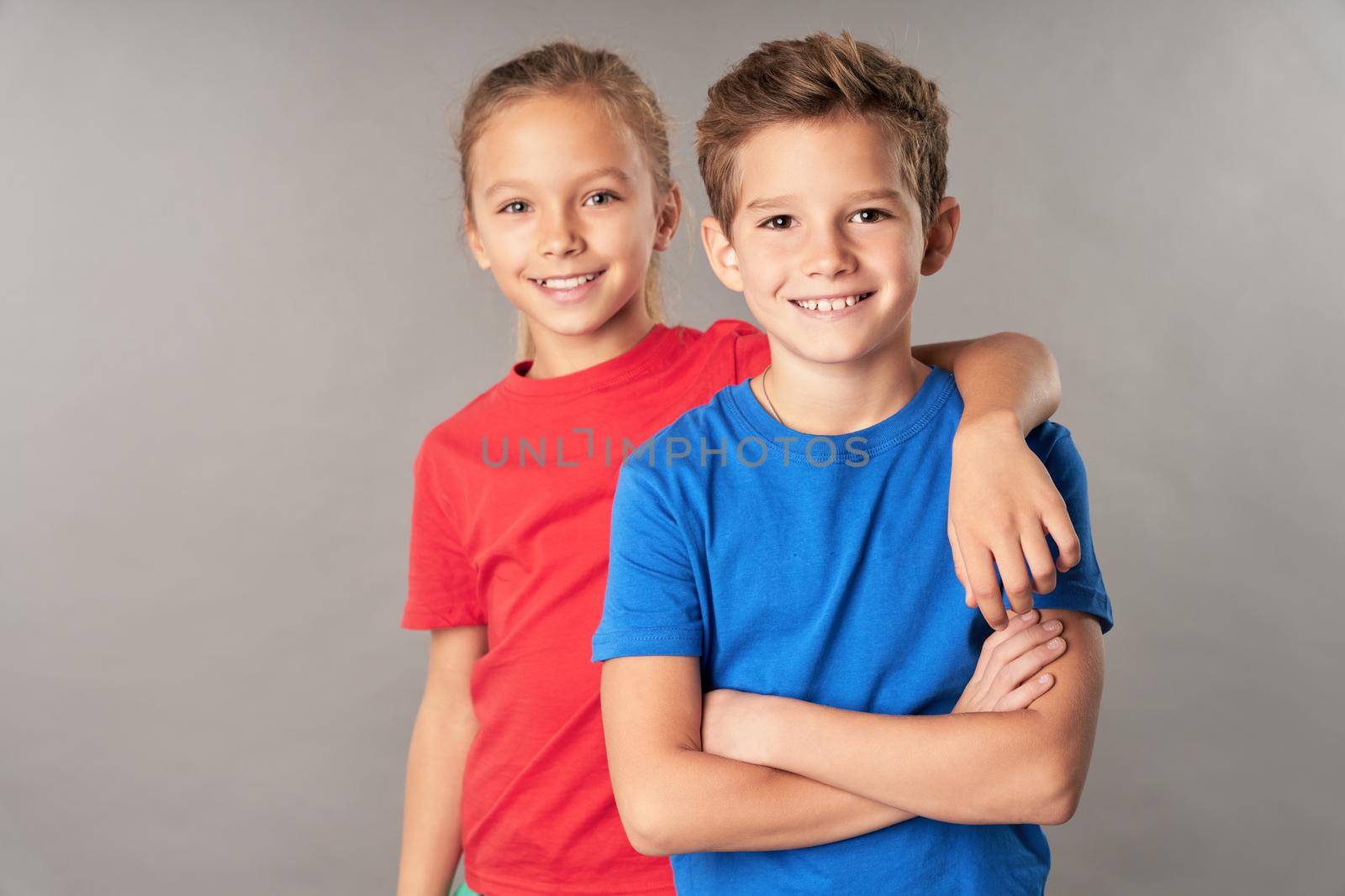 Adorable female child wrapping arm around brother shoulder and smiling while boy crossing arms over his chest