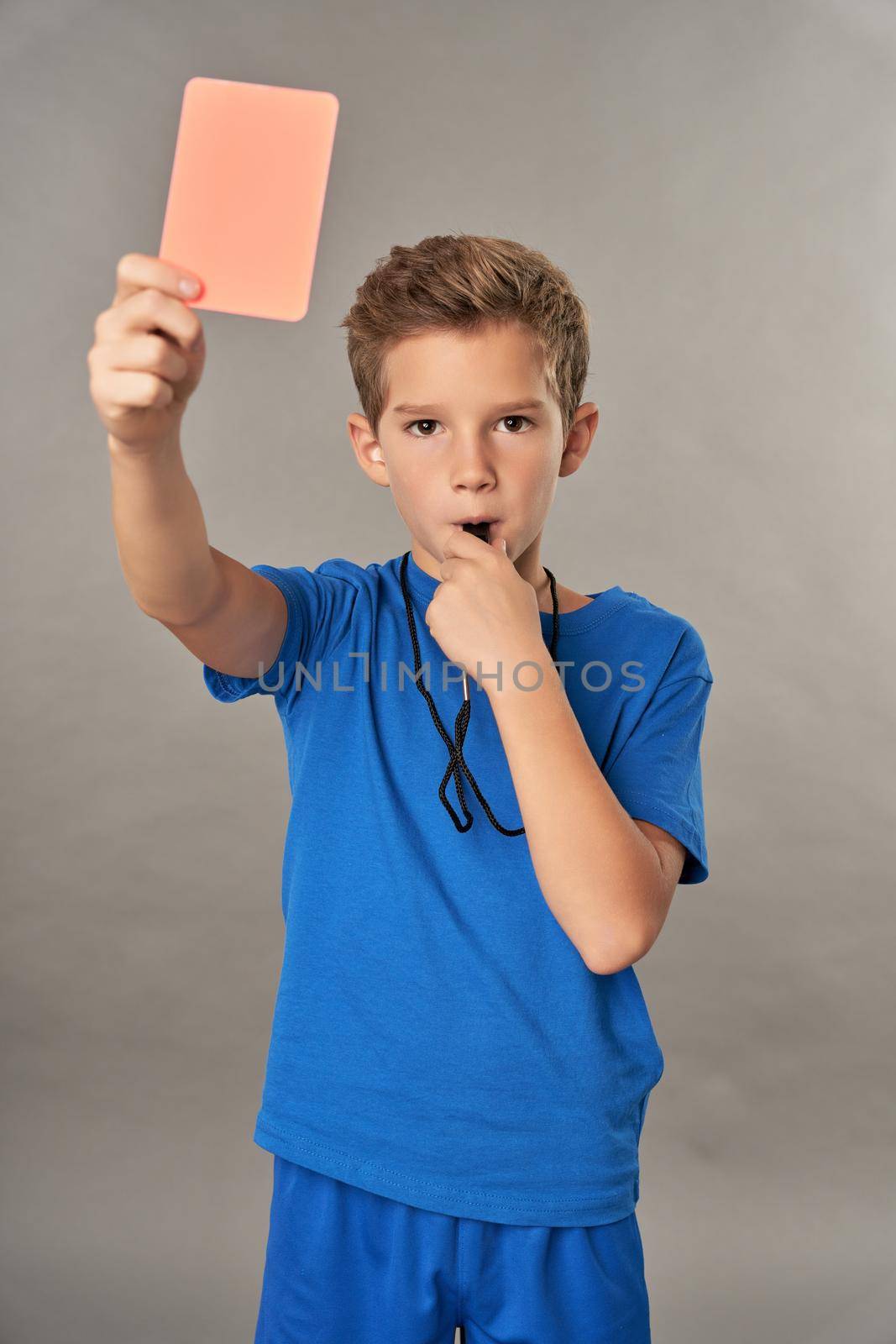 Adorable male kid using referee whistle and showing penalty card while standing against gray background