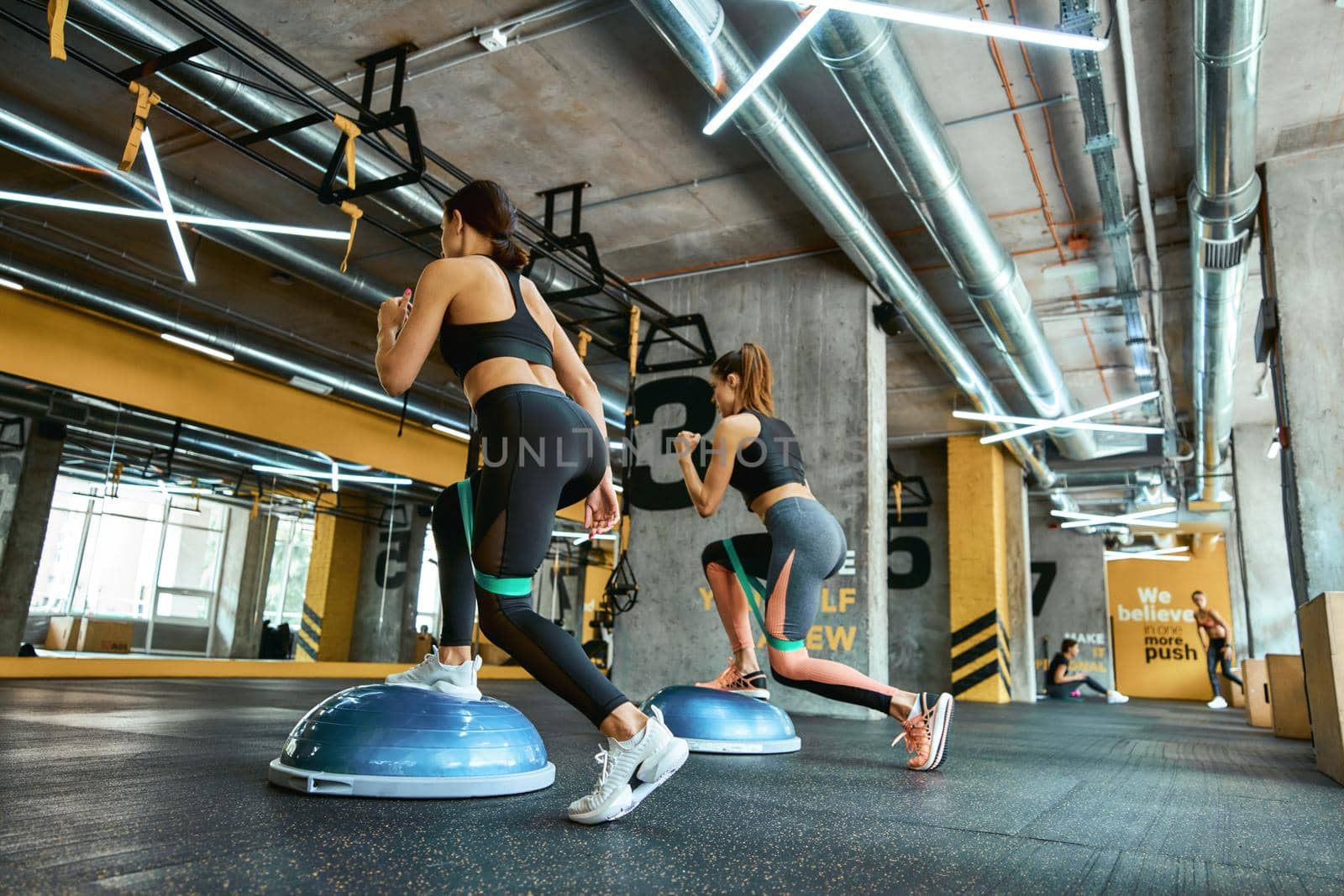 Full length of two young athletic women in sportswear exercising together with resistance fitness bands at crossfit gym, doing squats and training legs by friendsstock