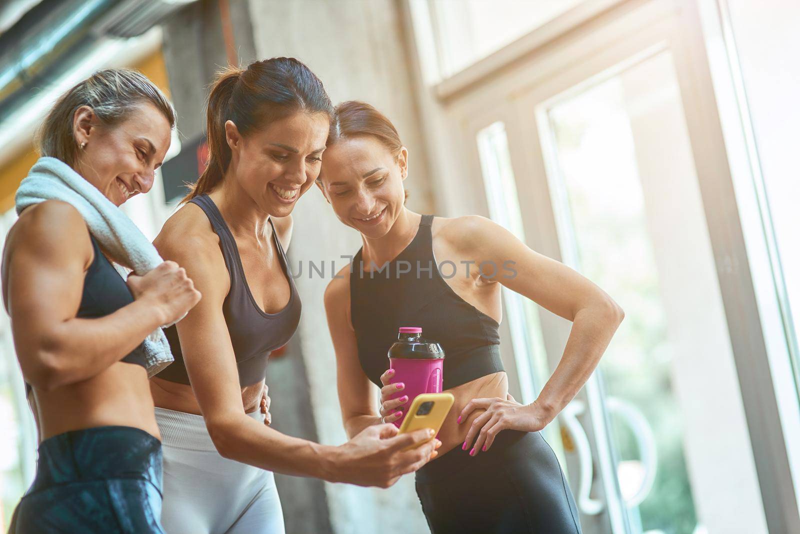 Group of three young beautiful and happy sportive women taking selfie on smartphone while resting after workout at gym, exercising together. Sport, wellness and healthy lifestyle