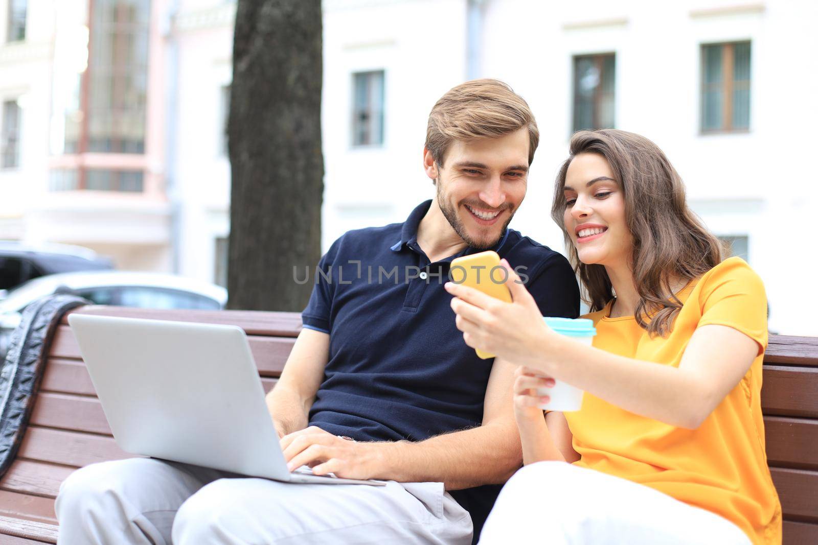 Attrative young couple using laptop computer while sitting on a bench outdoors, holding mobile phone