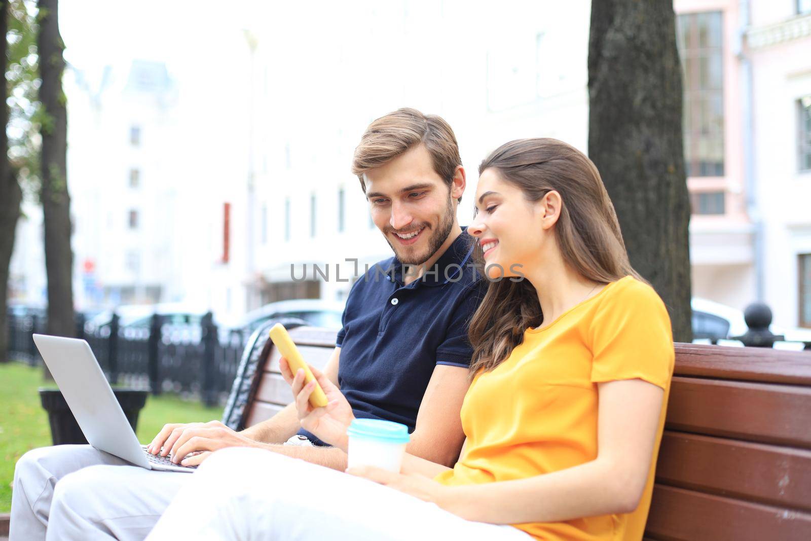Attrative young couple using laptop computer while sitting on a bench outdoors, holding mobile phone. by tsyhun