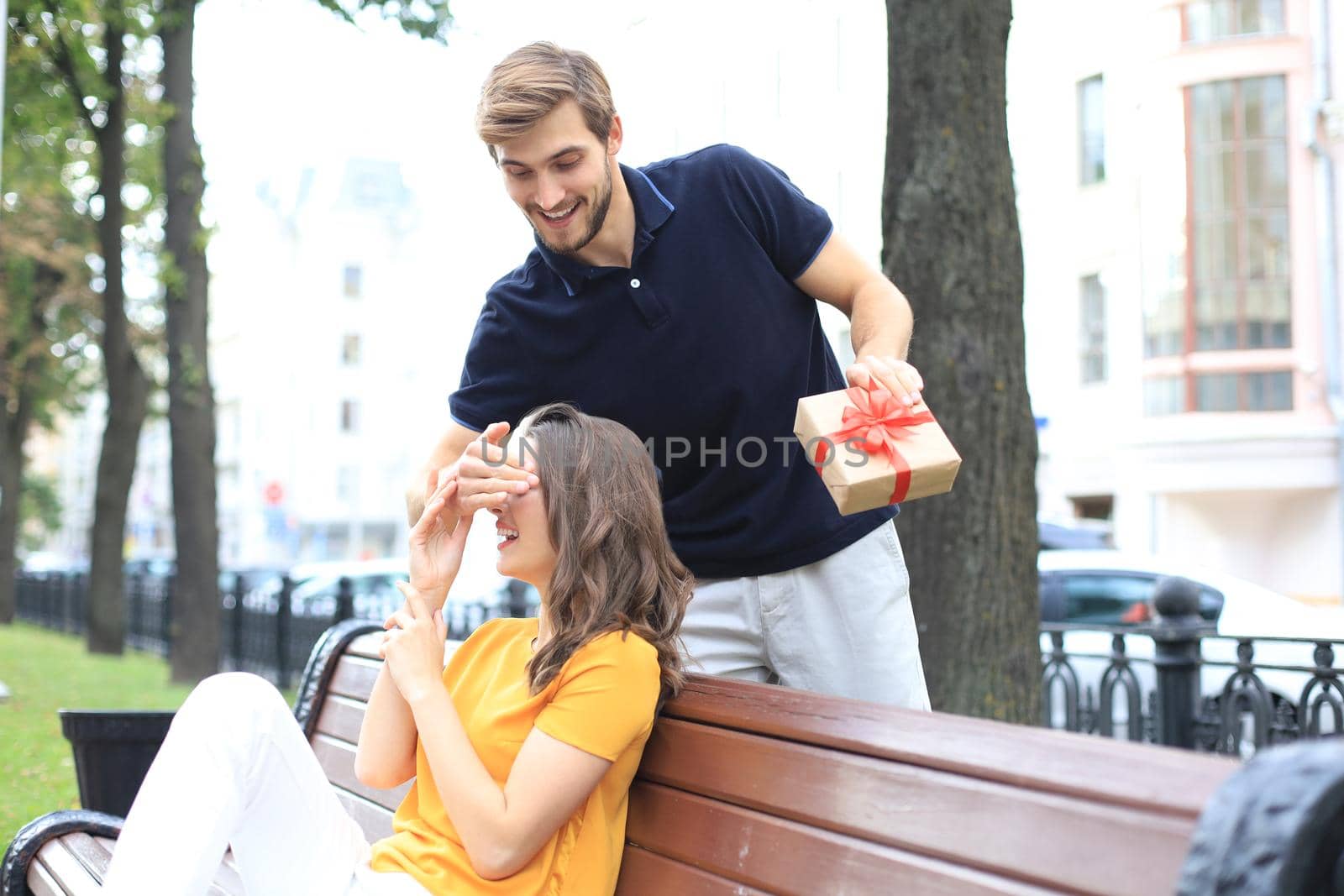 Image of charming excited couple in summer clothes smiling and holding present box together while sitting on bench