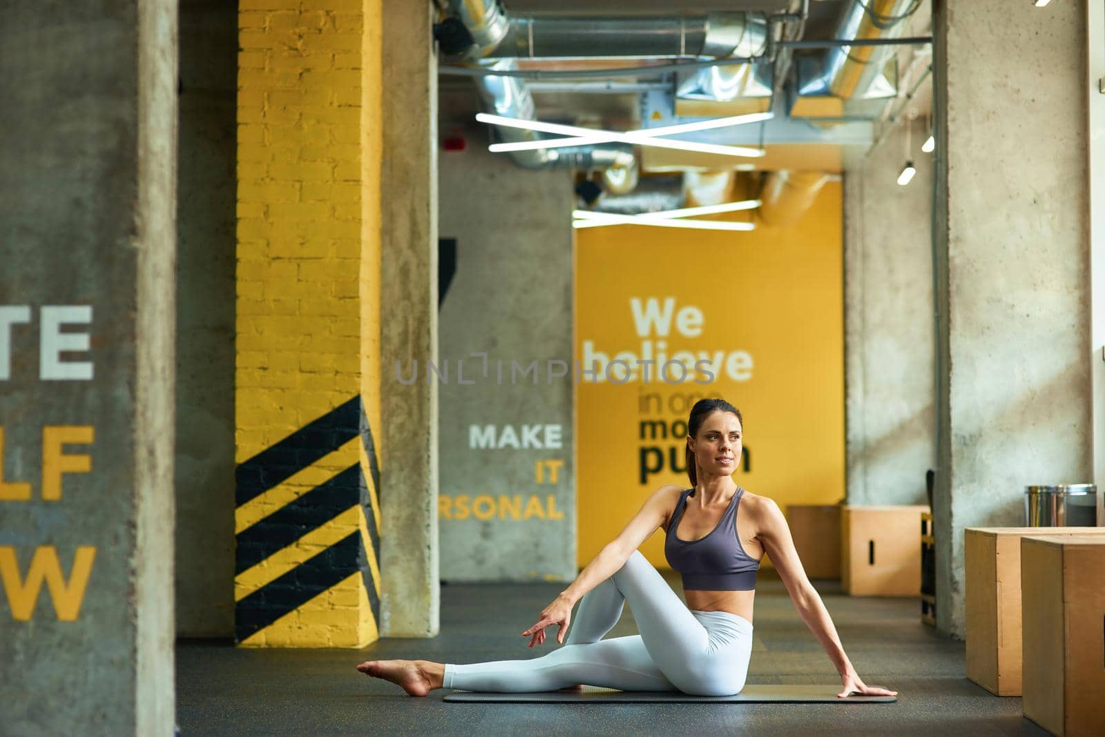 Full length of a young fitness woman in sportswear sitting on yoga mat at gym and warming up before workout, doing stretching exercises by friendsstock