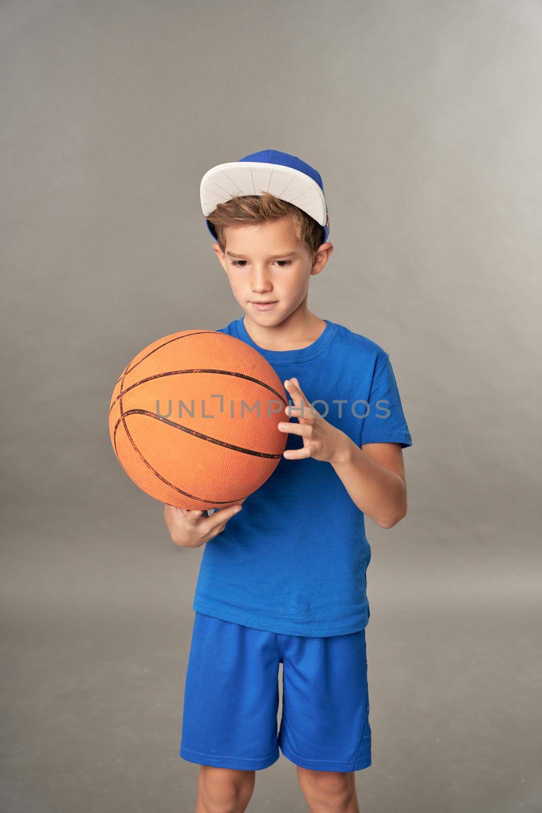 Cute male child in sportswear and cap looking at basketball ball in his hands while standing against gray background