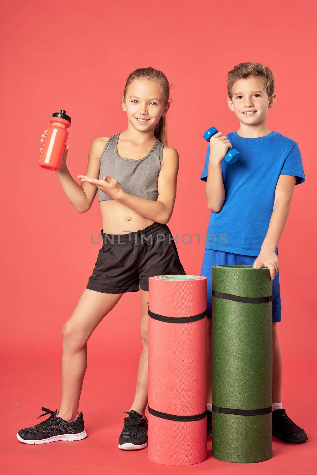 Adorable female child with bottle of water looking at camera and smiling while boy holding dumbbell and yoga mat