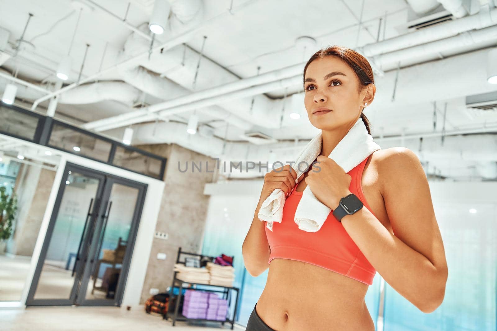 Resting after workout. Young beautiful fit woman with white towel on shoulders looking away while standing in fitness studio or gym. Sport, training, wellness and healthy lifestyle