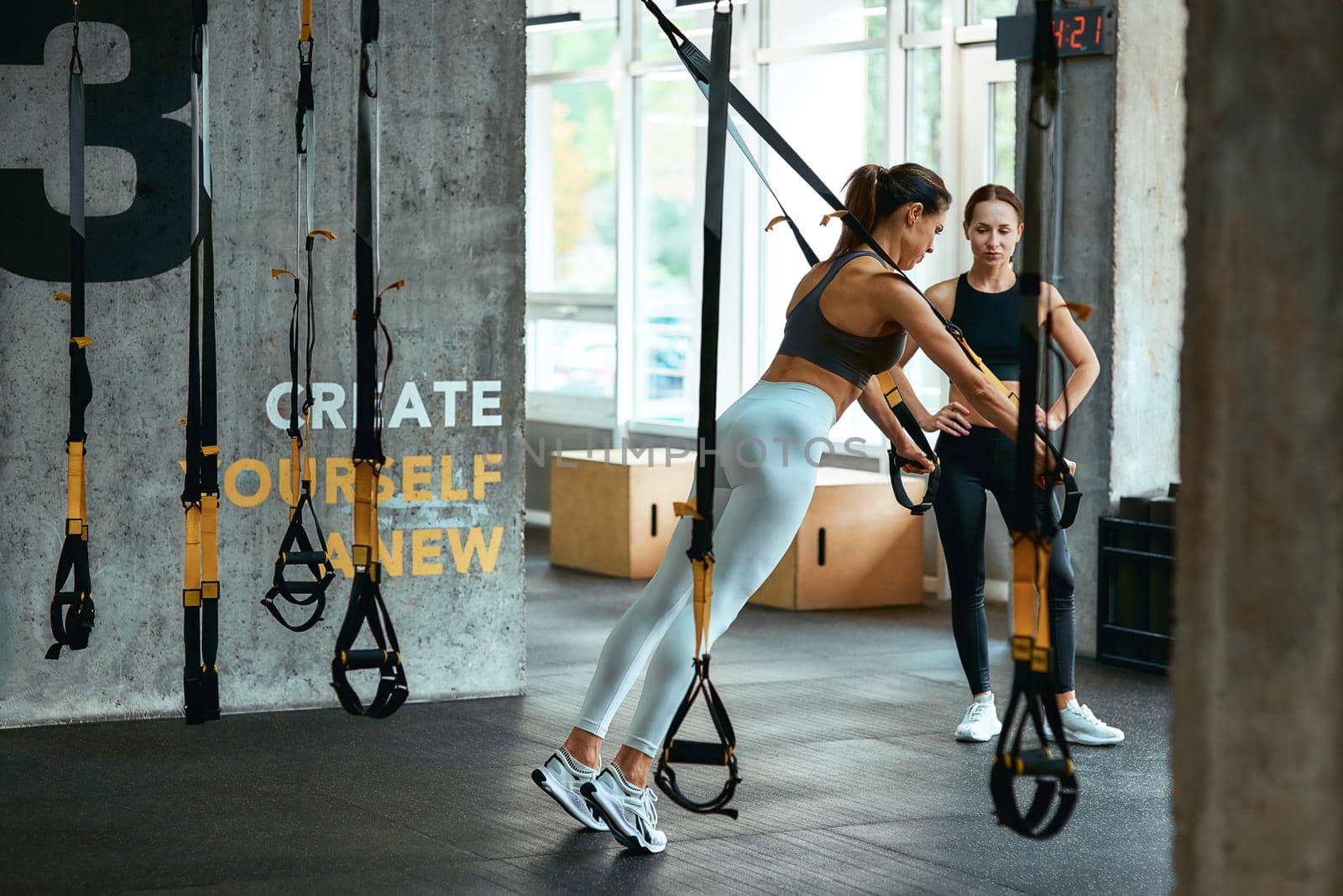 Young athletic woman in sportswear exercising with trx fitness straps with personal trainer at gym, doing push ups. Sport, training, wellness and healthy lifestyle