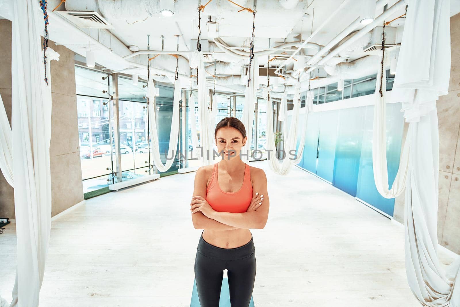 Young attractive and happy woman in sportswear, female fitness instructor keeping arms crossed and smiling at camera while standing in a beautiful fly yoga studio. Sport, wellness, healthy lifestyle