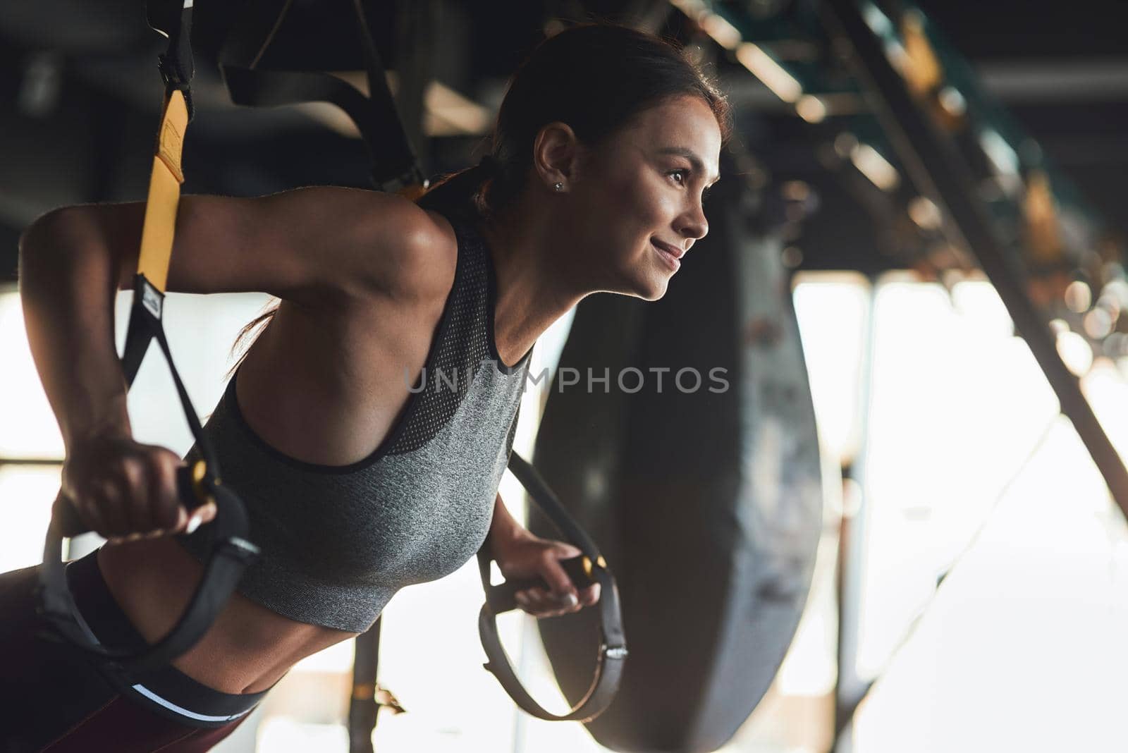 Side view of young beautiful athletic woman wearing sports clothes exercising with fitness trx straps at industrial gym, she is training arms and smiling by friendsstock