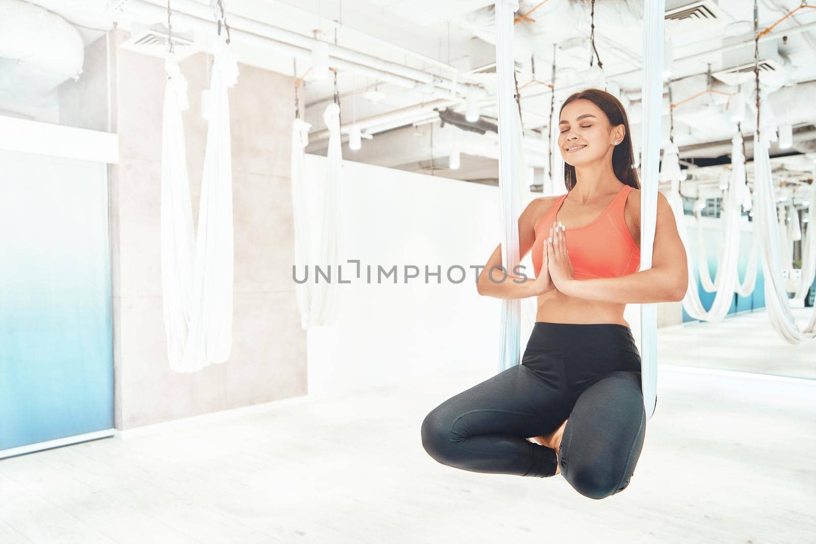 Young beautiful and happy fitness woman in sportswear sitting in lotus pose in white hanging hammock and meditating, doing fly yoga. Wellness and healthy lifestyle, harmony living