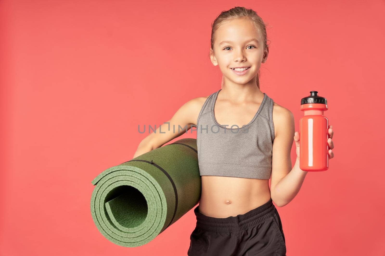 Smiling female child holding rolled exercise mat and refreshing sports drink while standing against red background