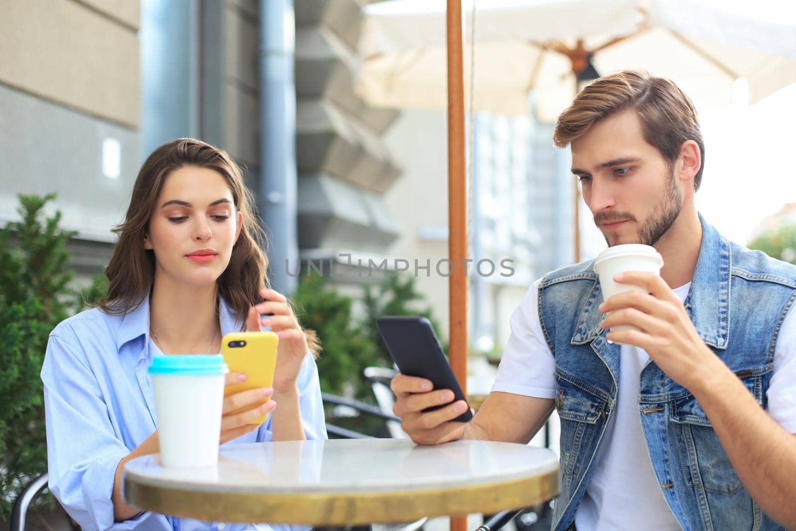 Attractive young couple in love drinking coffee while sitting at the cafe table outdoors, using mobile phone. by tsyhun