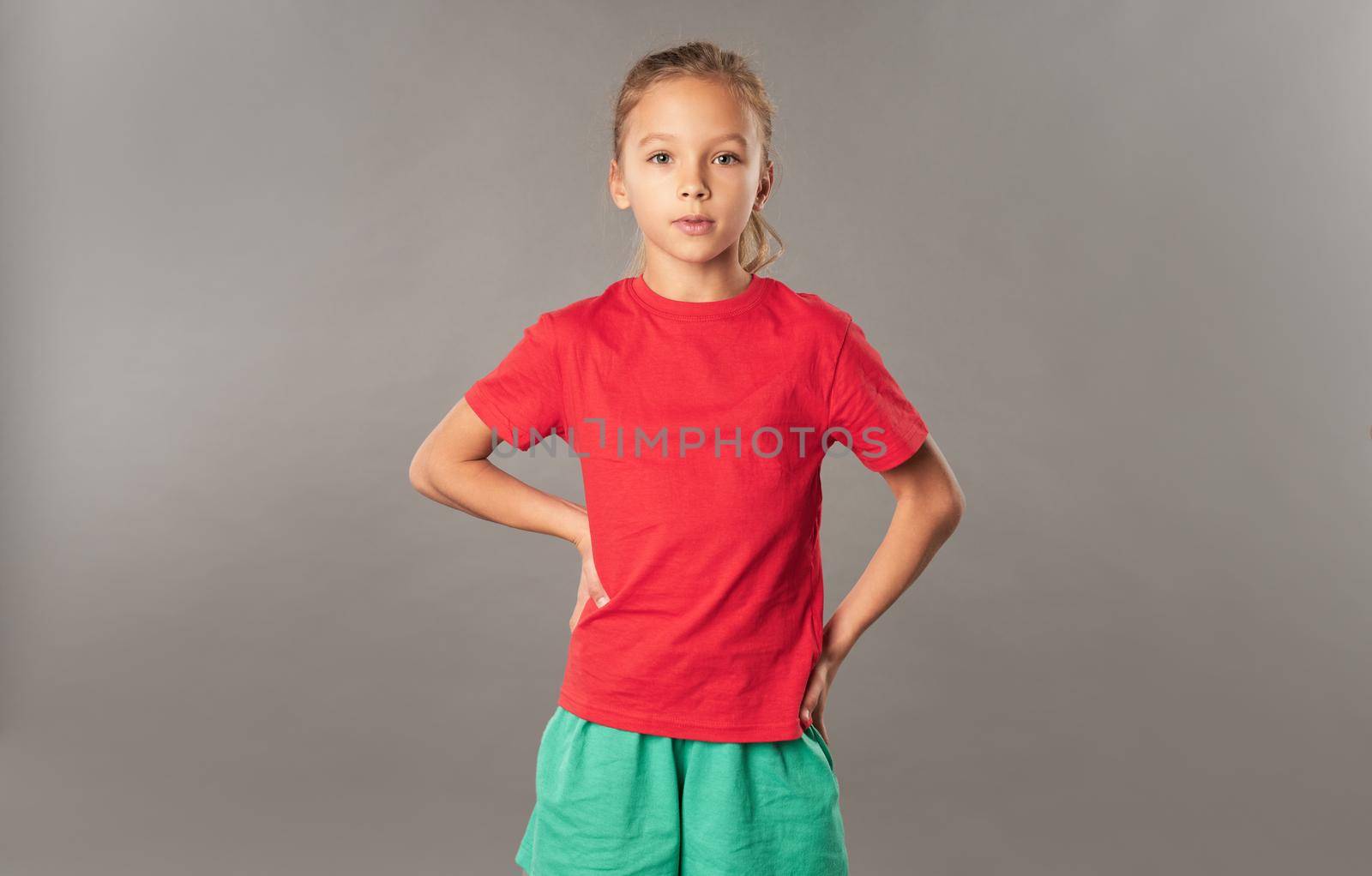 Adorable female child keeping hands on waist and looking at camera with serious expression