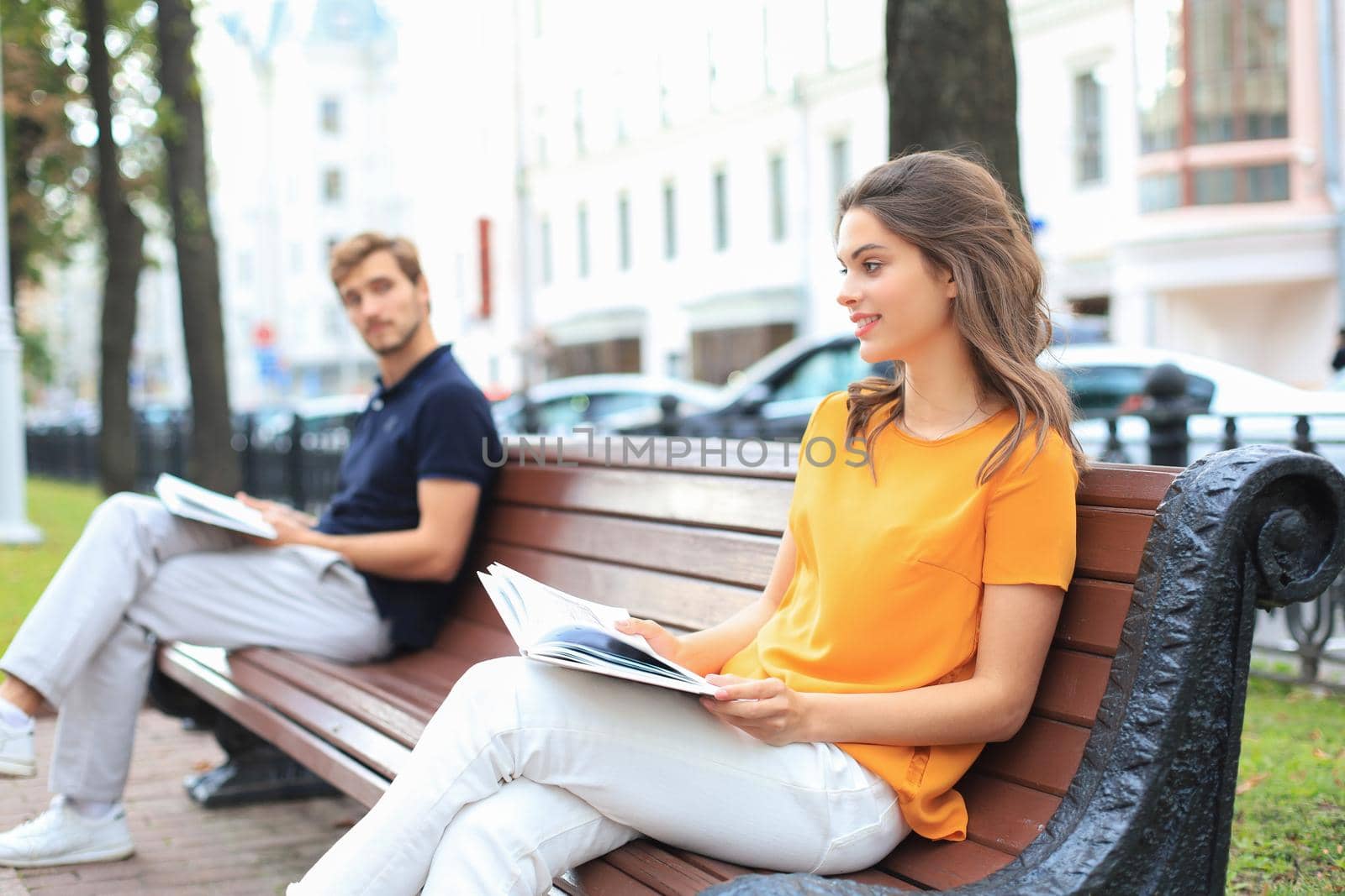Romantic young couple in summer clothes smiling and reading books together while sitting on bench in city street