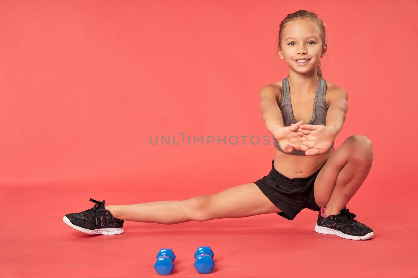 Adorable female child in sportswear stretching legs and smiling while working out with dumbbells. Isolated on red background