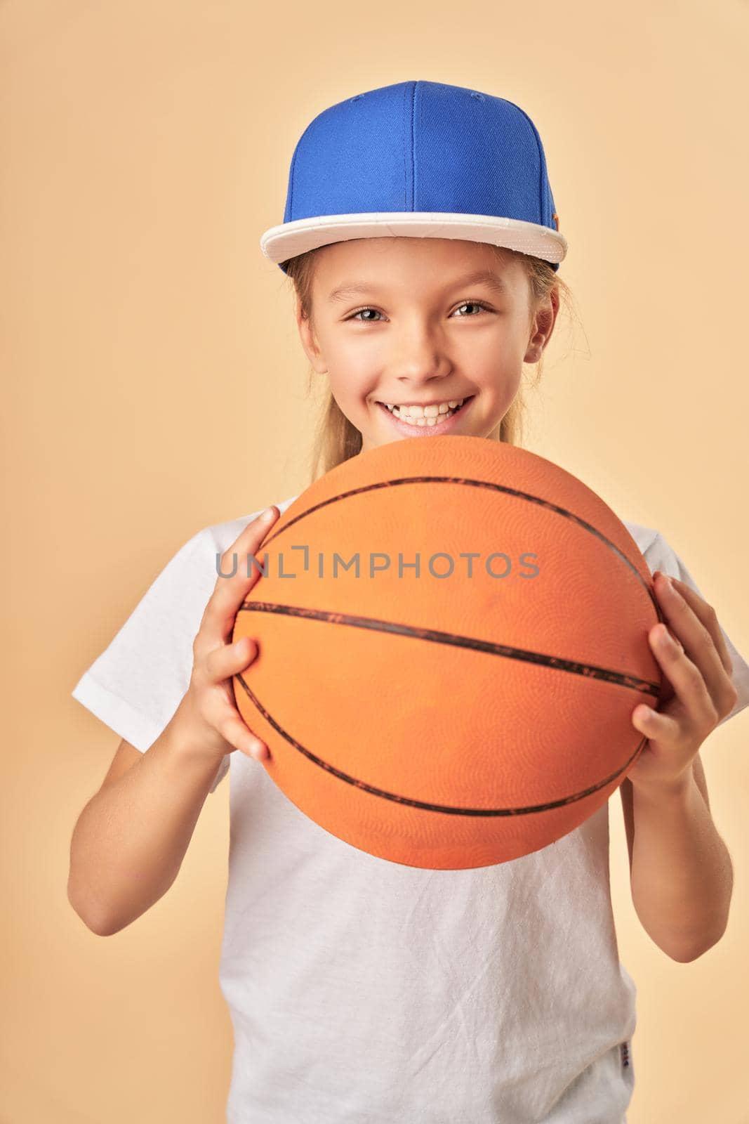Cute female child basketball player holding game ball and smiling while standing against light orange background