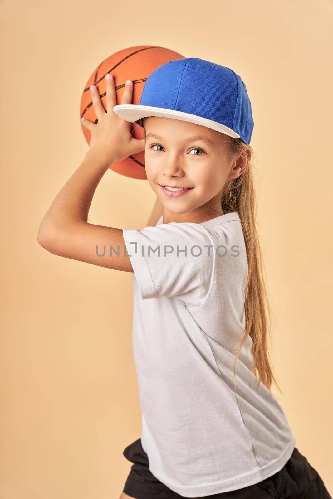 Cheerful female child basketball player holding game ball and smiling while standing against light orange background