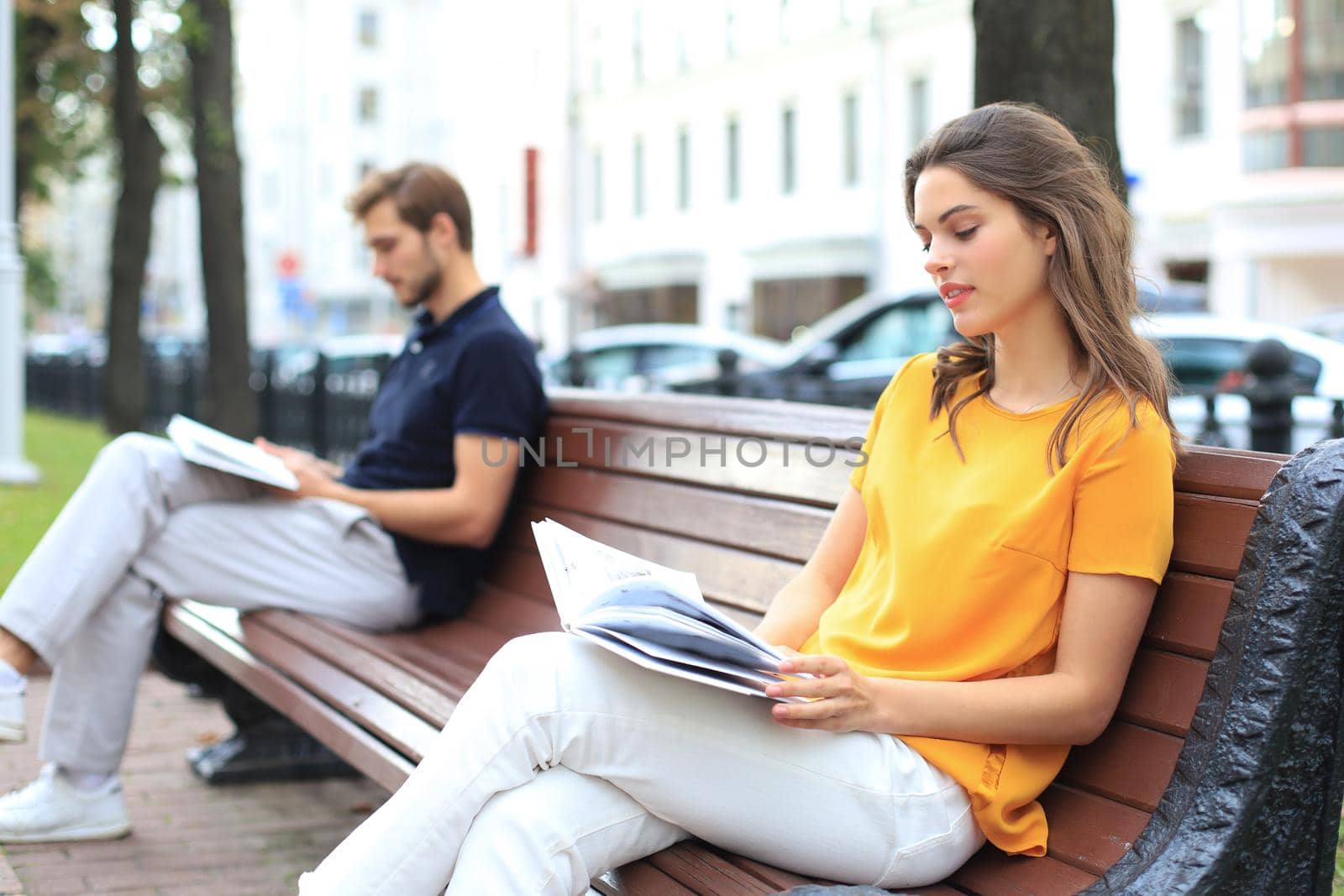 Romantic young couple in summer clothes smiling and reading books together while sitting on bench in city street