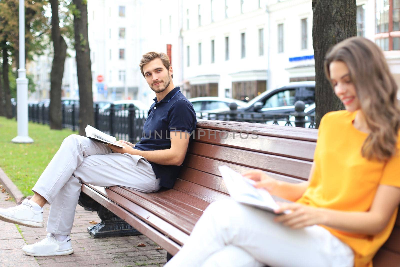 Romantic young couple in summer clothes smiling and reading books together while sitting on bench in city street. by tsyhun