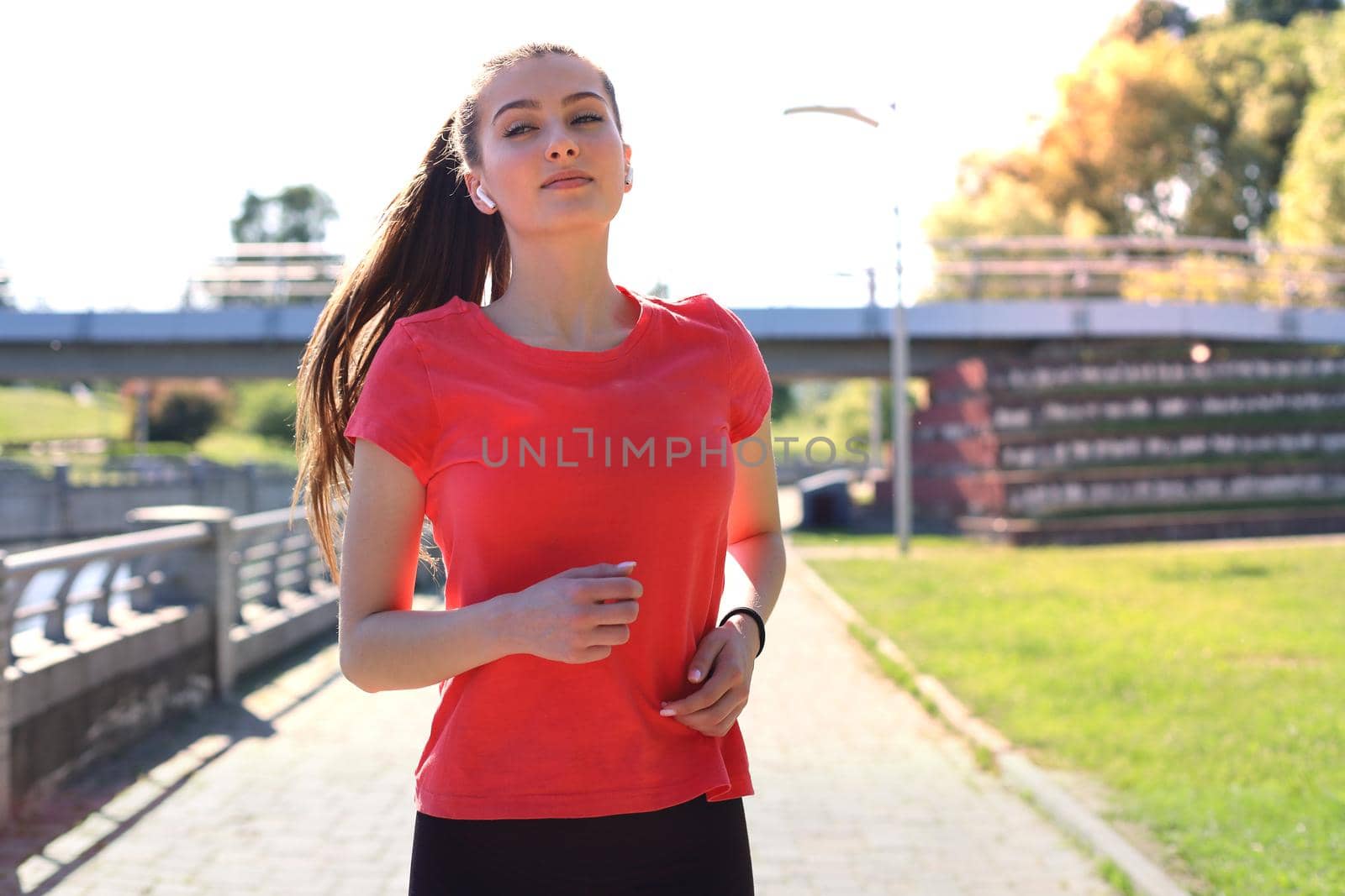Photo of sporty slim woman wearing sportswear listening music with earphones while running working out near city riverfront