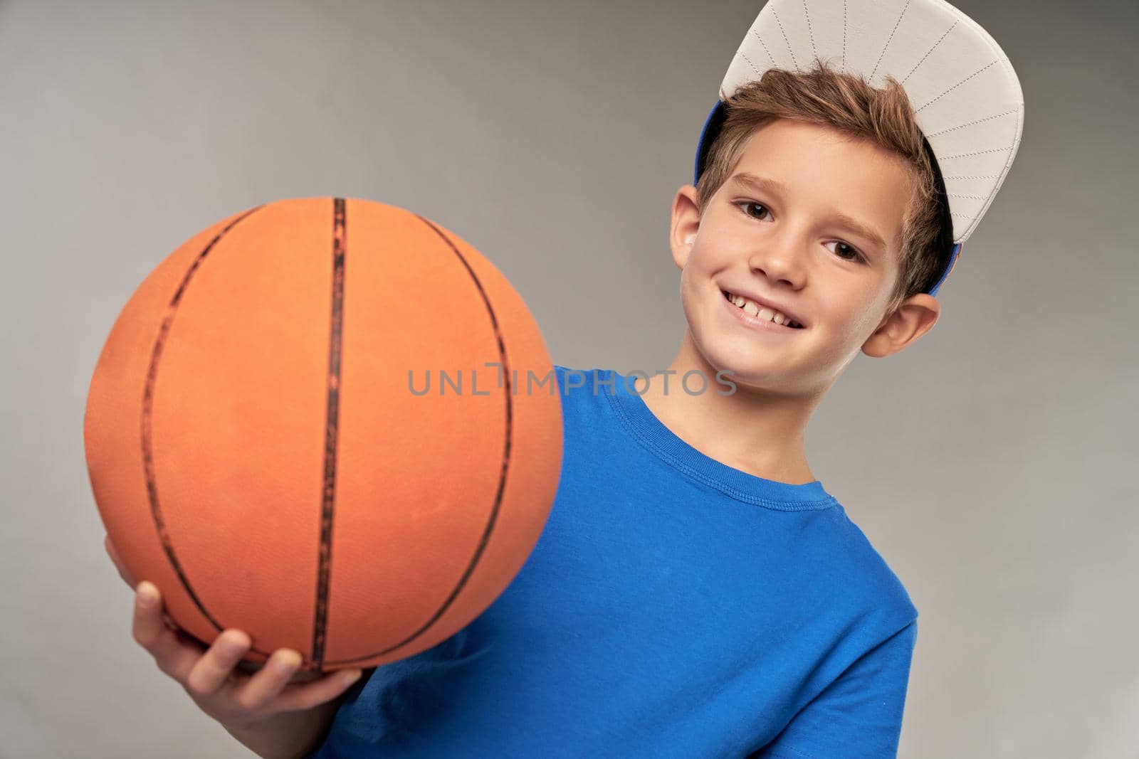 Cute boy basketball player holding game ball and smiling while standing against gray background