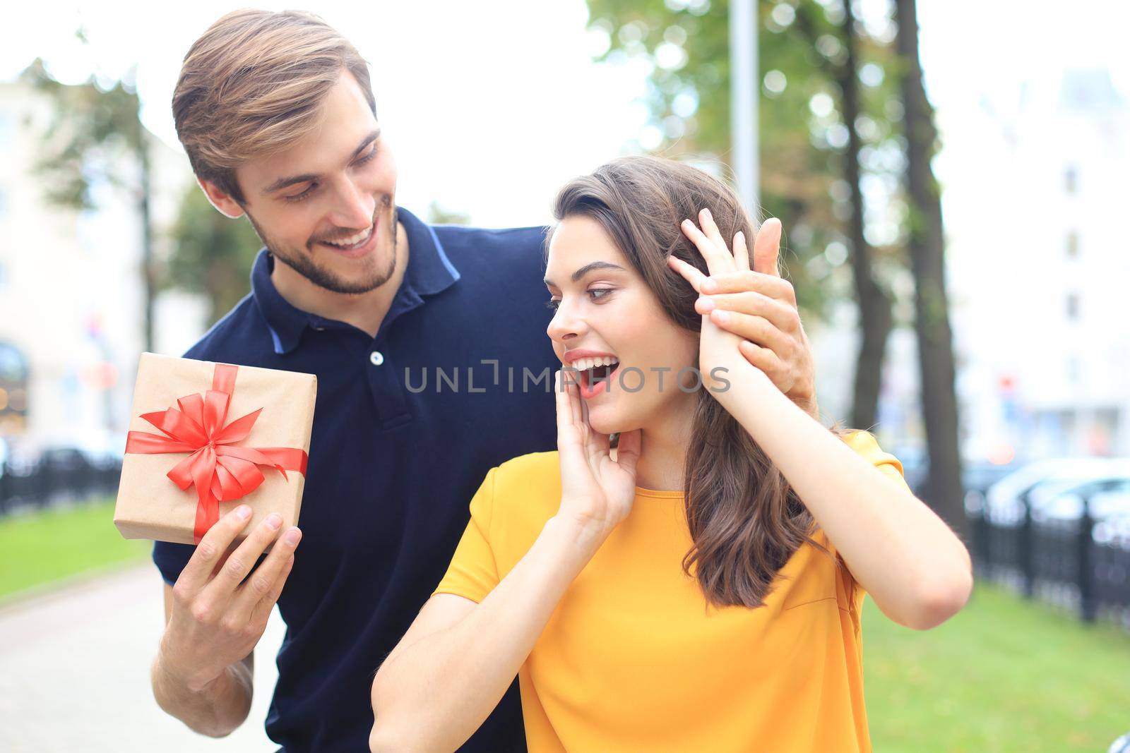 Astonished excited couple in summer clothes smiling and holding present box together while standing on city street