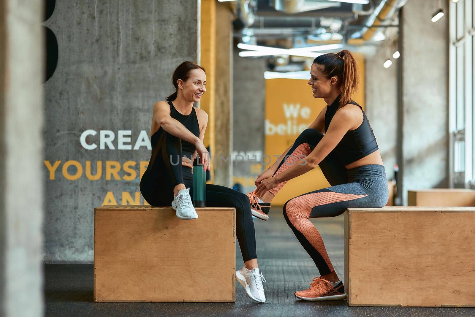 Full length of two young athletic fitness girls in sportswear sitting on crossfit jump boxes at gym and talking, exercising together. Sport, training, wellness and healthy lifestyle